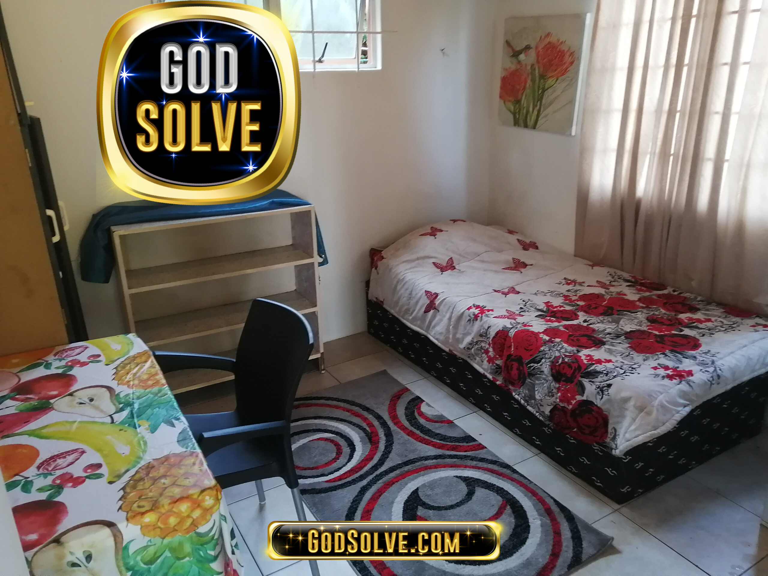 Small bedroom with a single bed covered in a red floral blanket, a black chair, desk with fruit-patterned cover, empty shelves, a window, and wall art. Logos read GOD SOLVE and GodSolve.com.